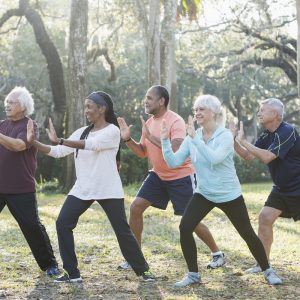 A group of five multi-ethnic seniors taking an exercise class in the park. They are practicing tai chi, standing with their hands raised.